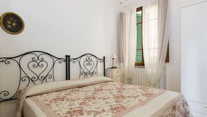 A bedroom with a bed, a nightstand, a lamp, a window with curtains, and a wall-mounted picture. - Belmare (Porto Azzurro)