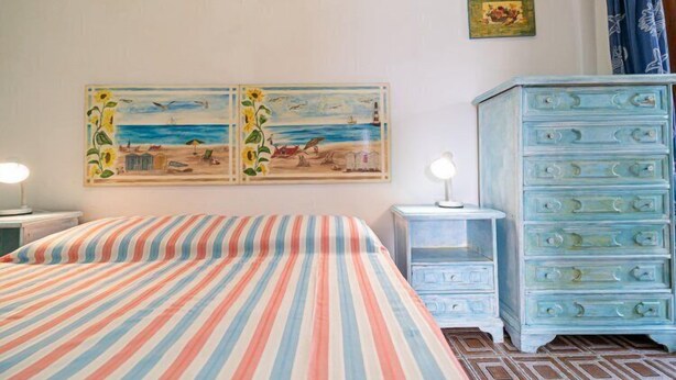 Um quarto com colcha listrada, duas pinturas de uma praia, cômoda azul e mesa de cabeceira com abajur.