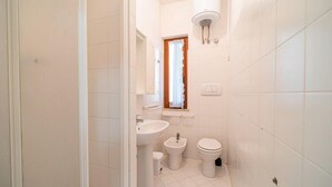 Uma casa de banho pequena e limpa, com azulejos brancos, uma pia, um bidé e uma sanita.