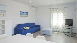 Quarto moderno e bem iluminado, com um sofá azul, um pequeno banquinho, uma televisão sobre um móvel e uma pintura com tema de praia na parede.