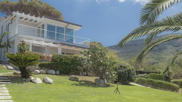 Uma casa moderna de dois andares com varanda, cercada por vegetação exuberante e um cenário montanhoso.