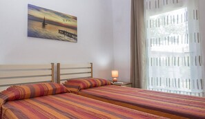 Quarto com duas camas, mesa de cabeceira com abajur, pintura de um pôr do sol sobre a água e janela com persianas.