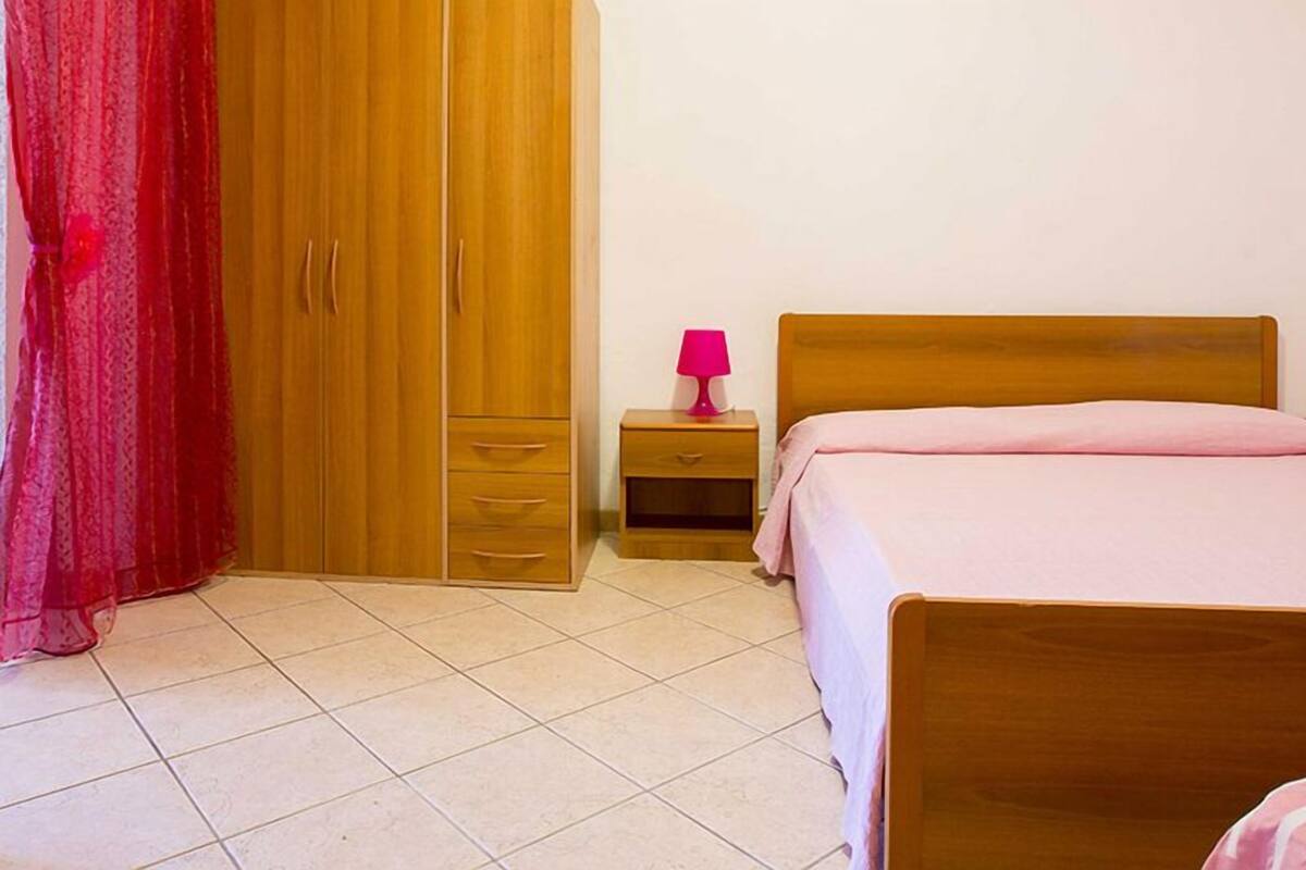 A bedroom with a wooden wardrobe, two bedside tables with lamps, a bed with a pink bedspread, and a red curtain.
