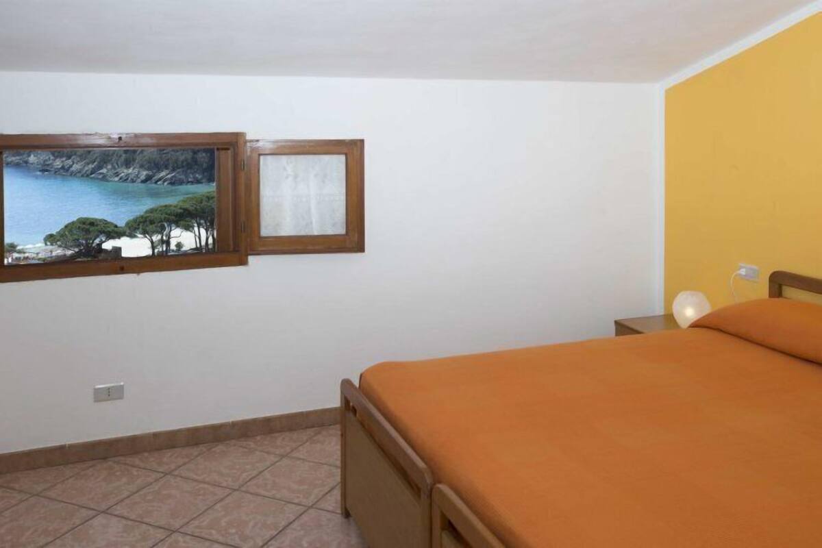 A bedroom with a bed, a nightstand, a lamp, and a window with a painting of a coastal scene.