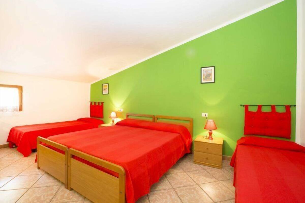 A room with three beds, each with a red bedspread, a wooden headboard, and a nightstand with a lamp. The walls are green, and there is a framed picture on the wall.