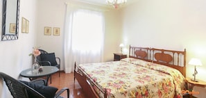 A bedroom with a wooden bed, a small round table, a chair, and a nightstand with a lamp. - Villa Medina (Porto Azzurro)