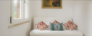 A white sofa with three patterned cushions, a window with a curtain, and a framed painting on the wall.