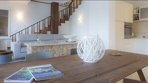 Een moderne woonkamer met een houten eettafel, een witte decoratieve vaas en een tijdschrift. Een trap leidt naar de bovenverdieping, en er is een keuken zichtbaar op de achtergrond.