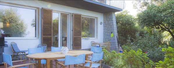 A patio with a wooden table and blue chairs, a stone wall, and a view of greenery.