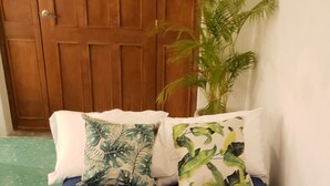 Una habitación con sofá cama azul, dos almohadas con motivos de hojas, una puerta de madera, una planta en maceta y un suelo con estampado verde.