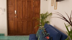 Una habitación con puerta de madera, un sofá azul con cojines estampados, un taburete de madera y una planta en maceta.