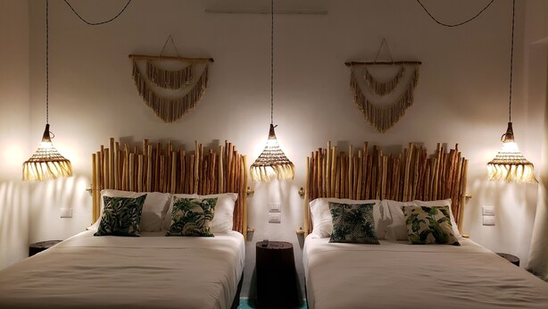 Una habitación con dos camas, cada una con ropa blanca y almohadas con estampado de hojas. Se observan decoraciones de macramé en las paredes y un cabecero de madera.