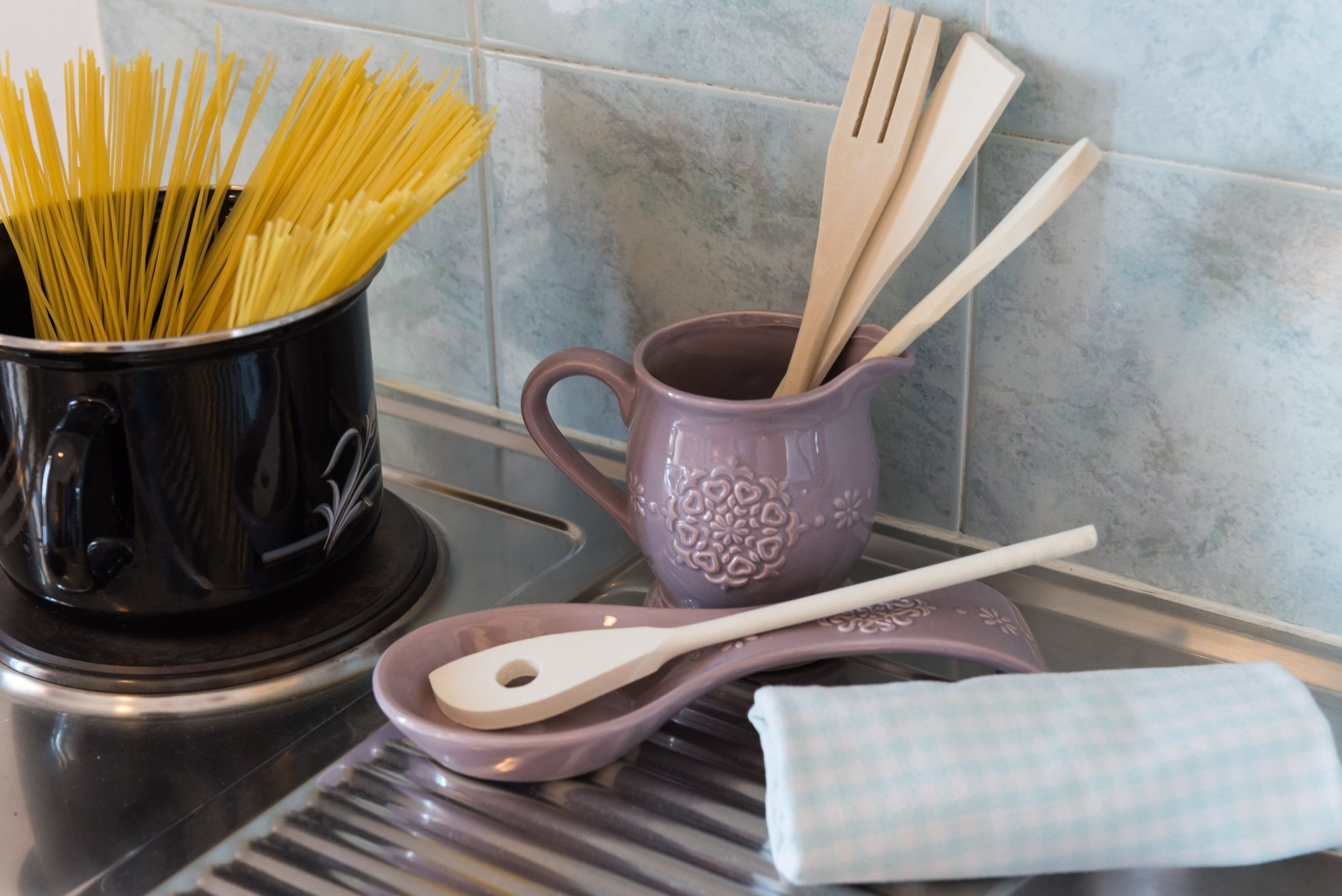 A kitchen counter with a pot of uncooked spaghetti, a purple mug, wooden utensils, and a checkered cloth.