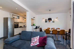 Een moderne woonkamer met een grijze bank, een eethoek met een houten tafel en stoelen, en een keuken met een koelkast en oven.