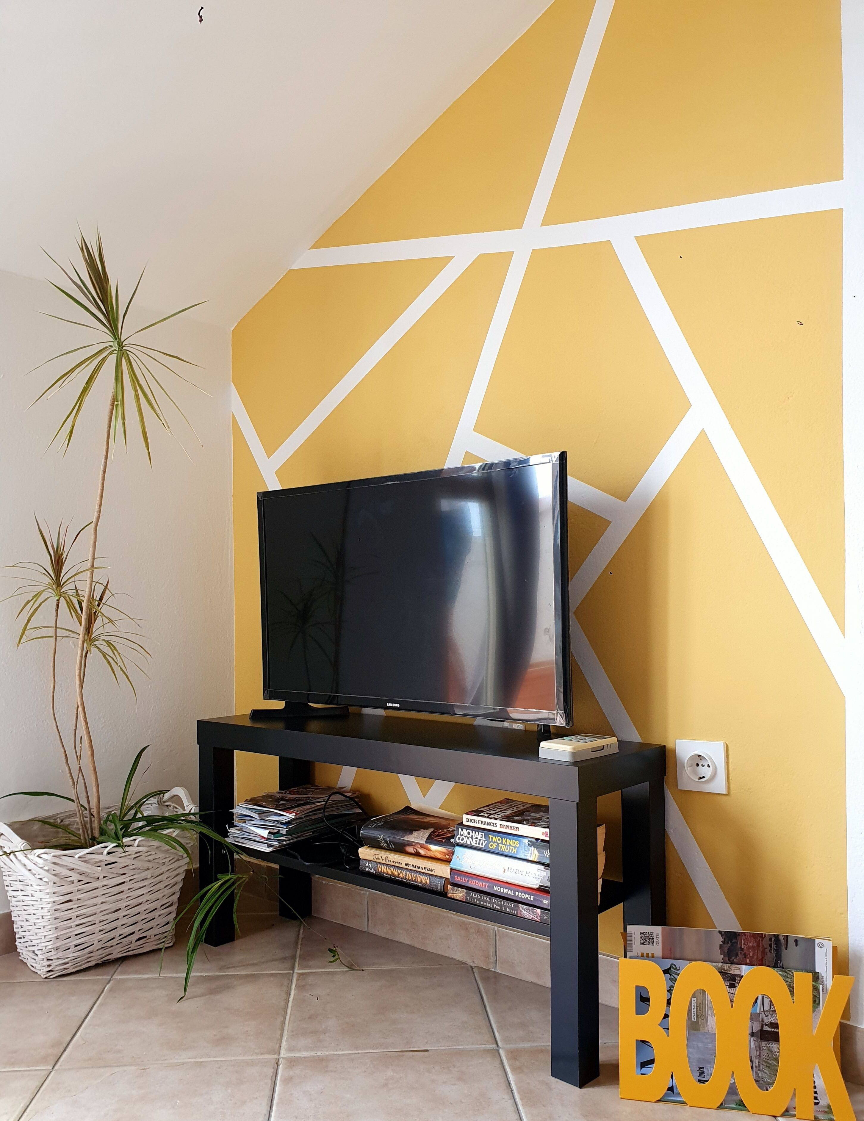 A living room with a TV on a black stand, a bookshelf with books, a potted plant, and a yellow wall with white geometric patterns.