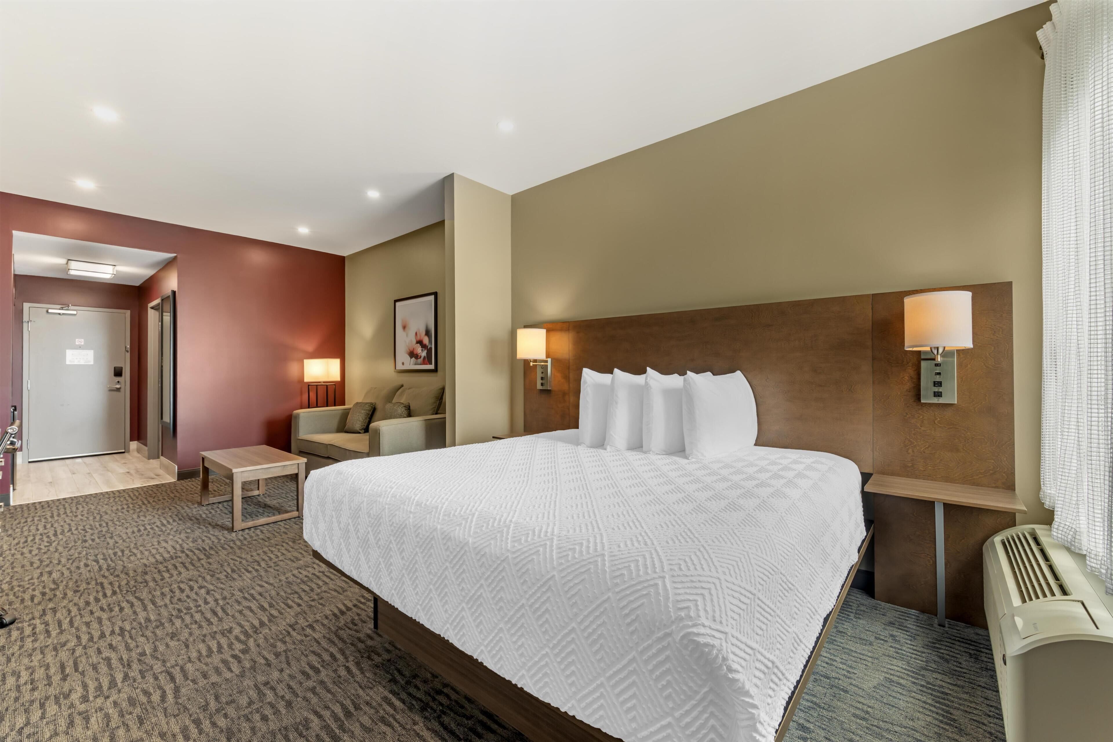 A hotel room with a large bed, a bedside table, a chair, a desk, and a wall-mounted lamp.