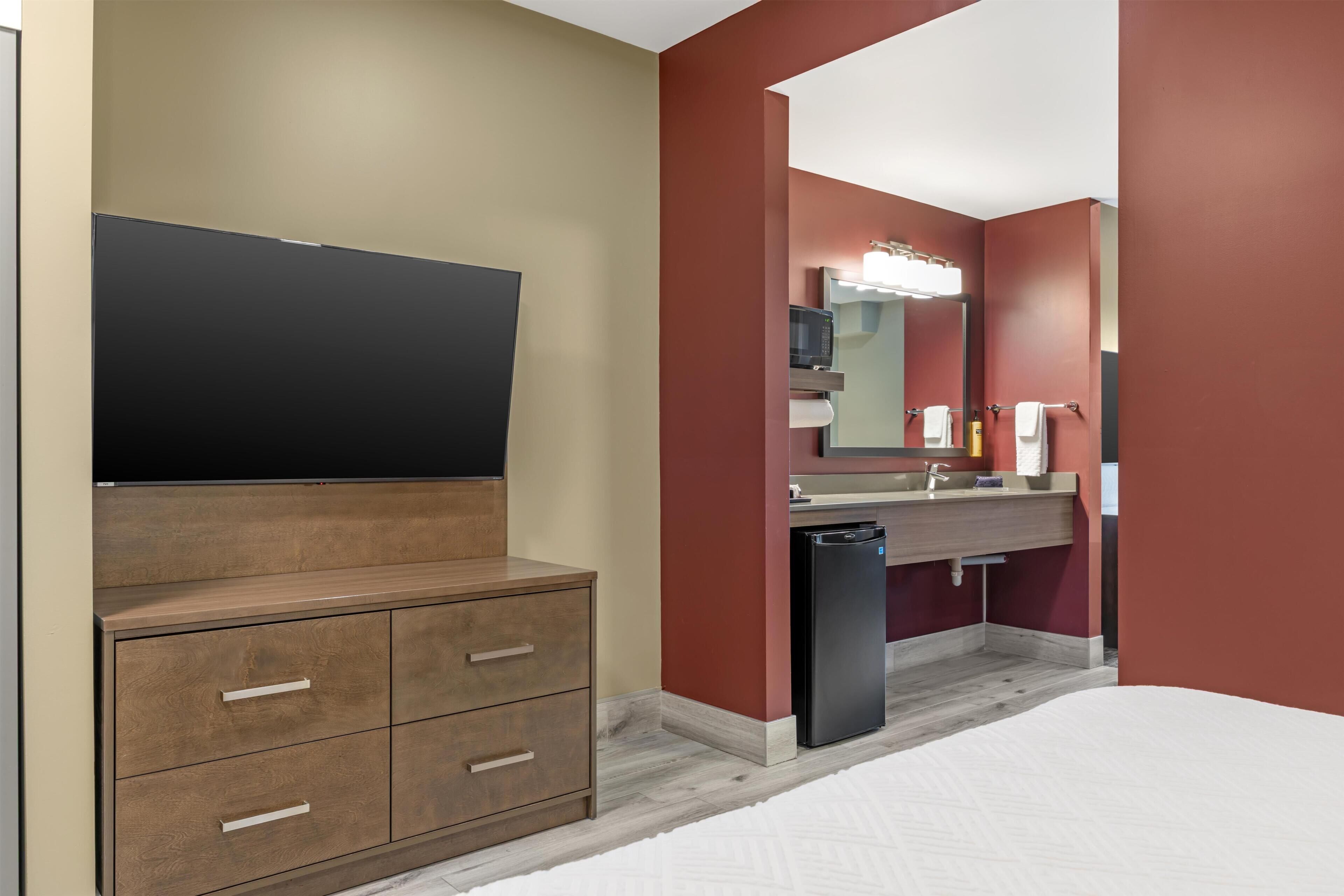 A hotel room with a wooden cabinet, a flat-screen TV, a sink with a mirror, and a compact refrigerator.