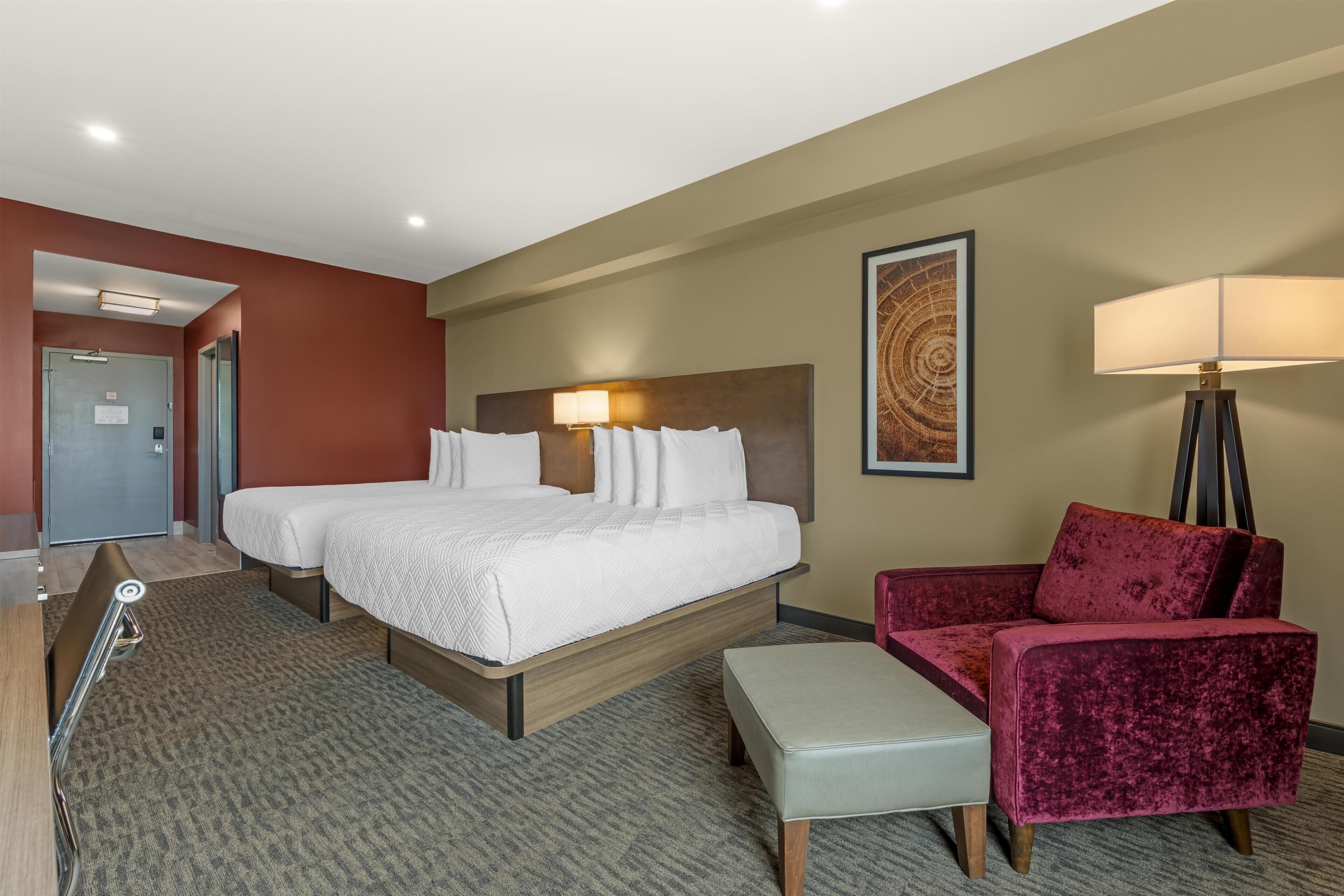 A hotel room with two beds, a desk, a chair, a lamp, and a painting on the wall.