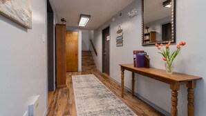 A hallway with a wooden floor, a rug, a wooden console table with a vase of flowers, a mirror, and a staircase in the background.