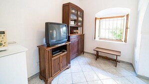 Een kamer met een televisie op een houten standaard, een kast met glazen deuren, een bankje en een raam met tralies.