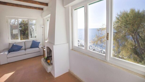 A room with a white sofa, a stone fireplace, and large windows with a view of the sea and trees. - Villa Casa Bianca (Campo nell'Elba)