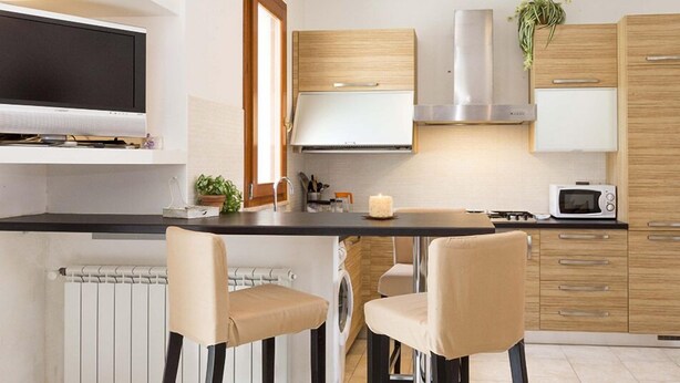 A modern kitchen with a flat-screen TV, a microwave, and wooden cabinets. There are two chairs at the dining table and a plant on the countertop. - Giulia (Campo nell'Elba)