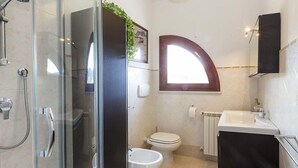 A bathroom with a glass shower enclosure, a toilet, a sink, and a window with a curved design. - Giulia (Campo nell'Elba)