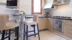 A modern kitchen with a gas stove, oven, and a TV mounted above the counter. There are two bar stools with cushioned seats. - Giulia (Campo nell'Elba)