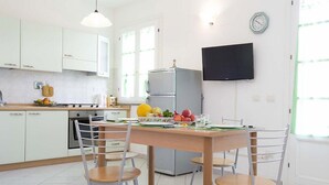 A modern kitchen with white cabinets, a wooden dining table, and chairs. There is a refrigerator, a stove, and a television mounted on the wall.