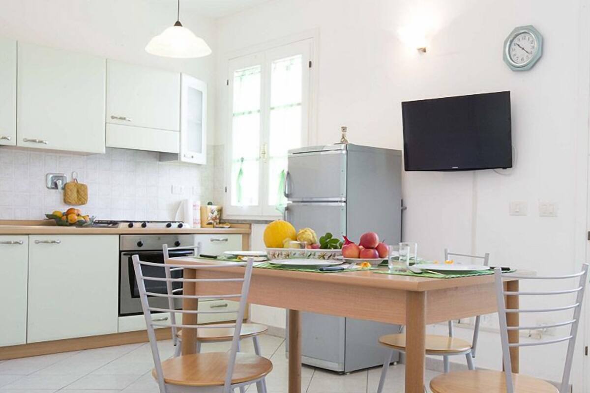 Une cuisine moderne avec des meubles blancs, une table à manger en bois et des chaises. On y trouve un réfrigérateur, une cuisinière et un téléviseur fixé au mur.
