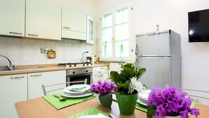 A kitchen with white cabinets, a stove, a sink, and a refrigerator. There are green placemats on the table and purple flowers in pots.