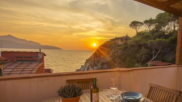 A table with a bottle of wine, glasses, and a potted plant on a balcony overlooking the sea at sunset. - App.ti Scaglieri Mare 1° piano (Portoferraio)