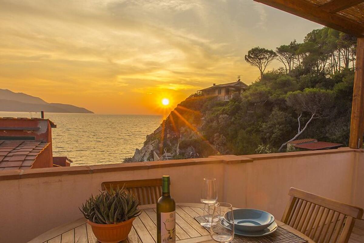A table with a bottle of wine, glasses, and a potted plant on a balcony overlooking the sea at sunset.