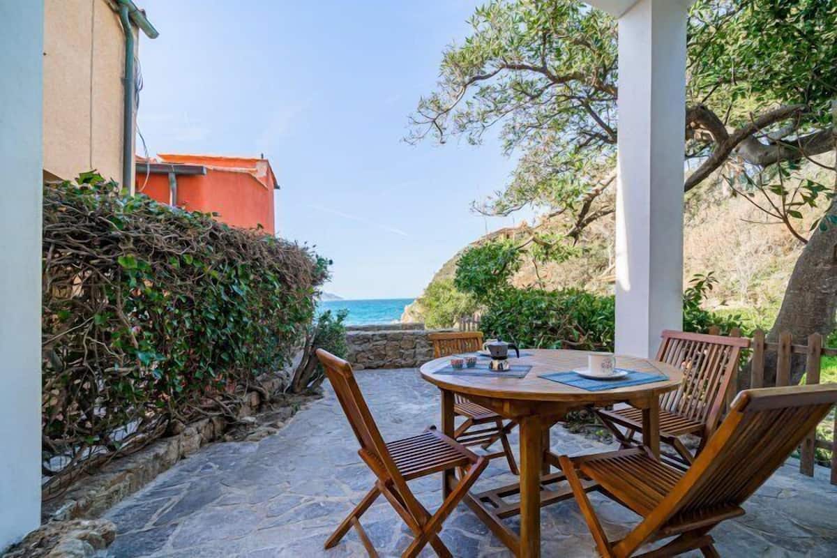 A patio with a round wooden table and chairs, overlooking a coastal area with a stone wall and vegetation.