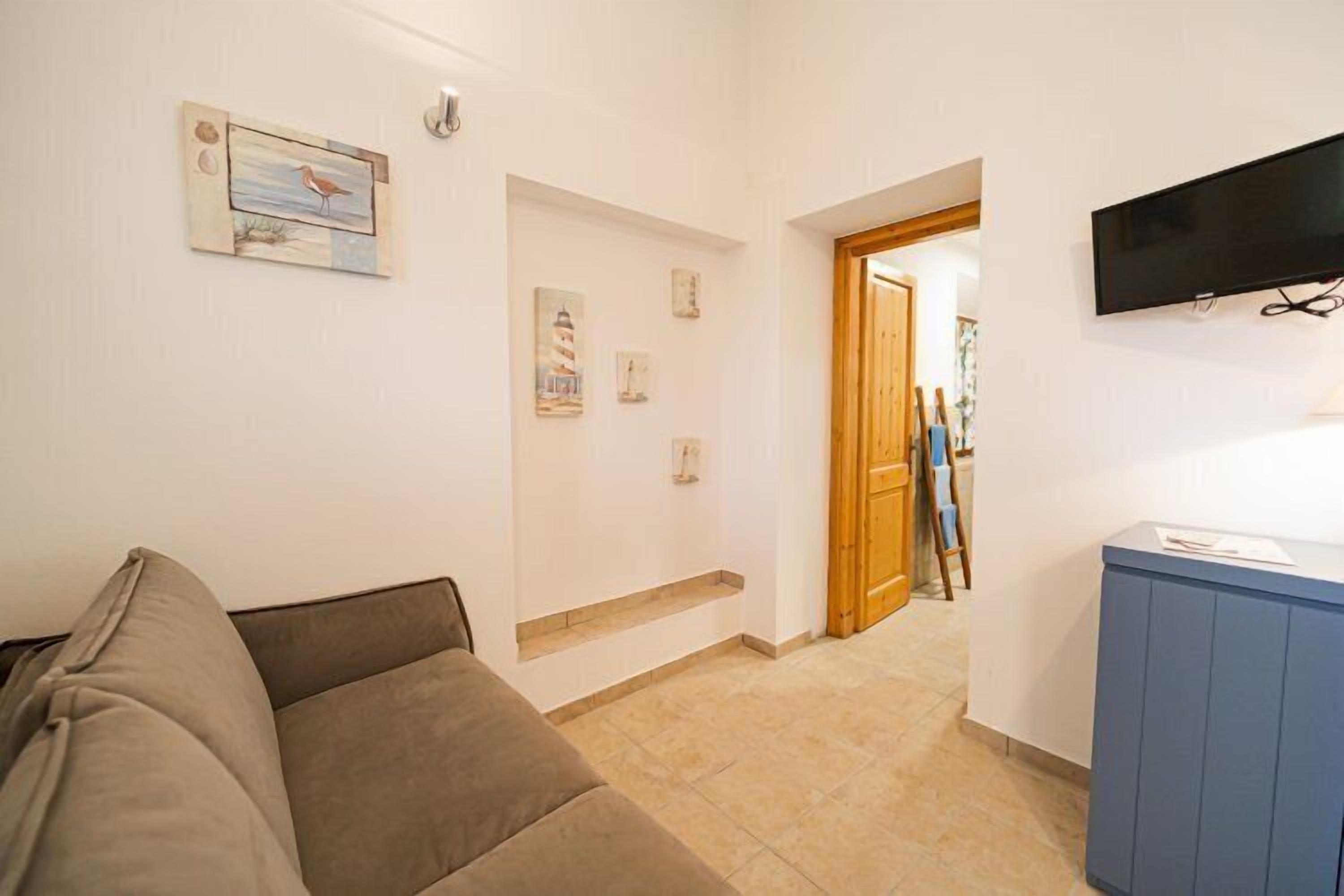 A living room with a sofa, a flat-screen TV mounted on the wall, and a wooden door leading to another room.