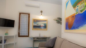 A living room with a beige sofa, a flat-screen TV, a painting of a sailboat, and a small desk with a lamp. - App.ti Scaglieri Mare 1° piano (Portoferraio)