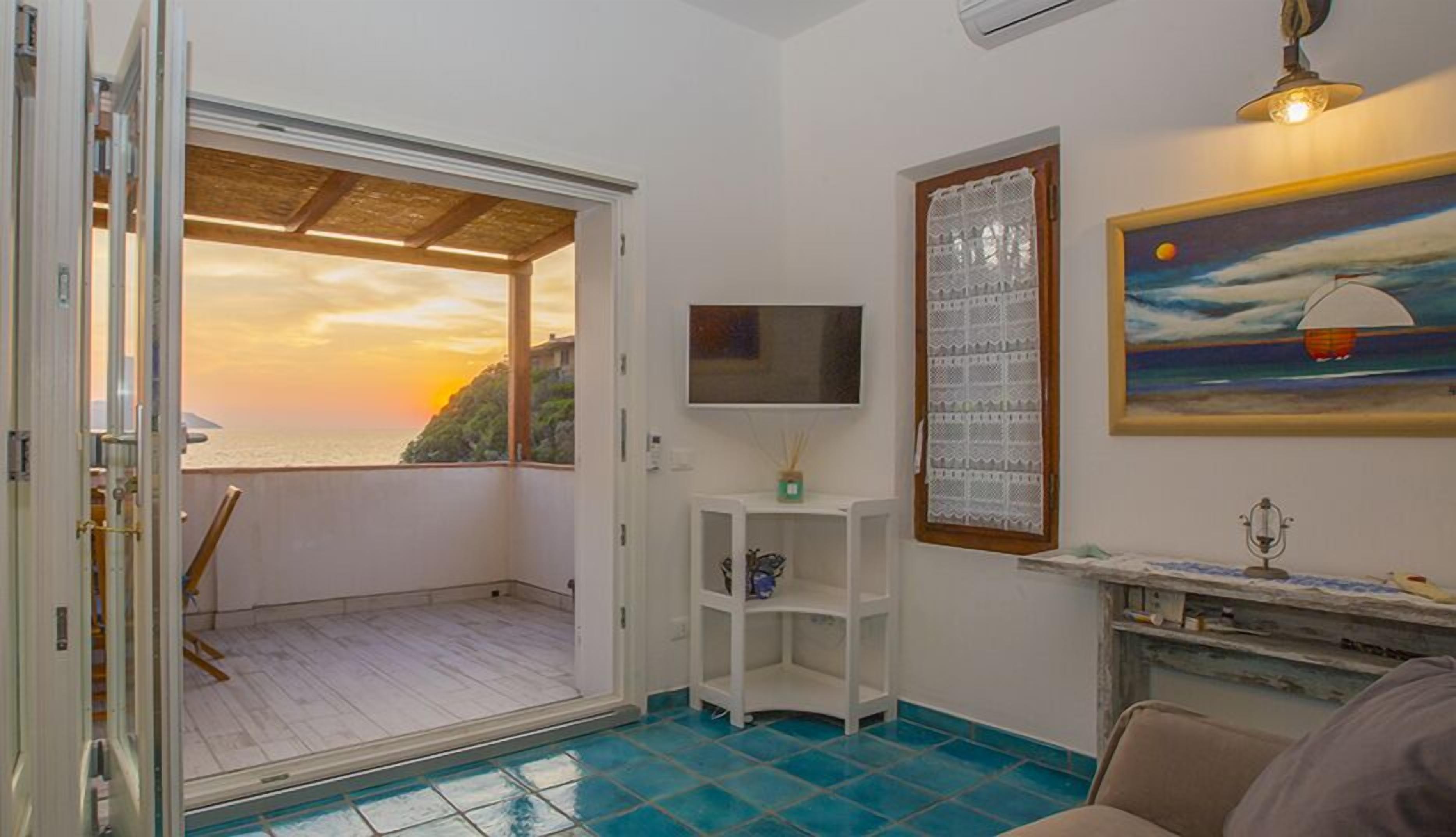 Habitación con vista al mar al atardecer, televisión, un cuadro y un pequeño estante blanco.