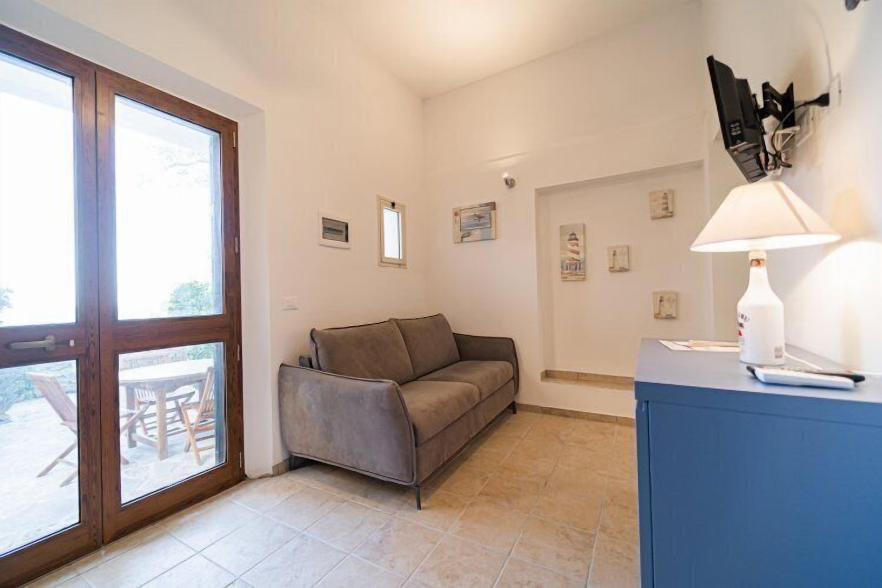 Una habitación con sofá, escritorio con lámpara y ventana con vista a muebles de exterior.