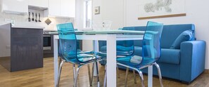 A modern kitchen with white cabinets, a glass dining table, and blue chairs. - Nila (Porto Azzurro)