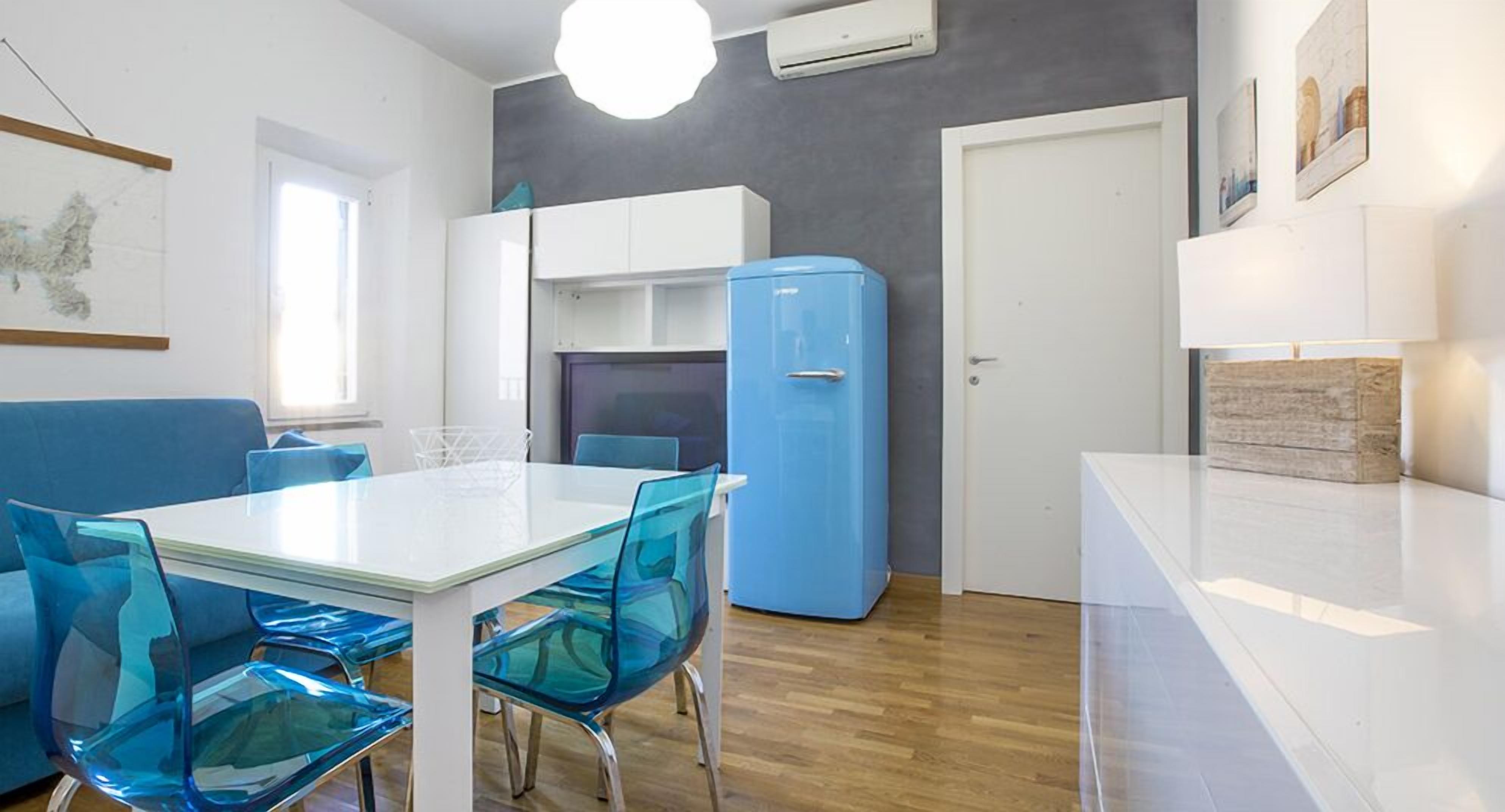 Une cuisine moderne avec une table blanche, des chaises bleues, un réfrigérateur bleu et des meubles de rangement blancs.
