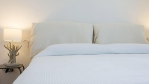 Una cama bien hecha con sábanas y almohadas blancas, una lámpara de noche con base decorativa, y una lámpara de pared.