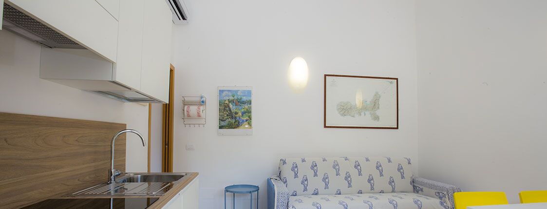 A small kitchenette with a sink and stove, a sofa with patterned cushions, and two framed pictures on the wall.