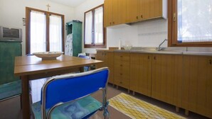A small, simple kitchen with wooden cabinets, a sink, and a dining table with a blue chair. - Santa Petronilla (Capoliveri)