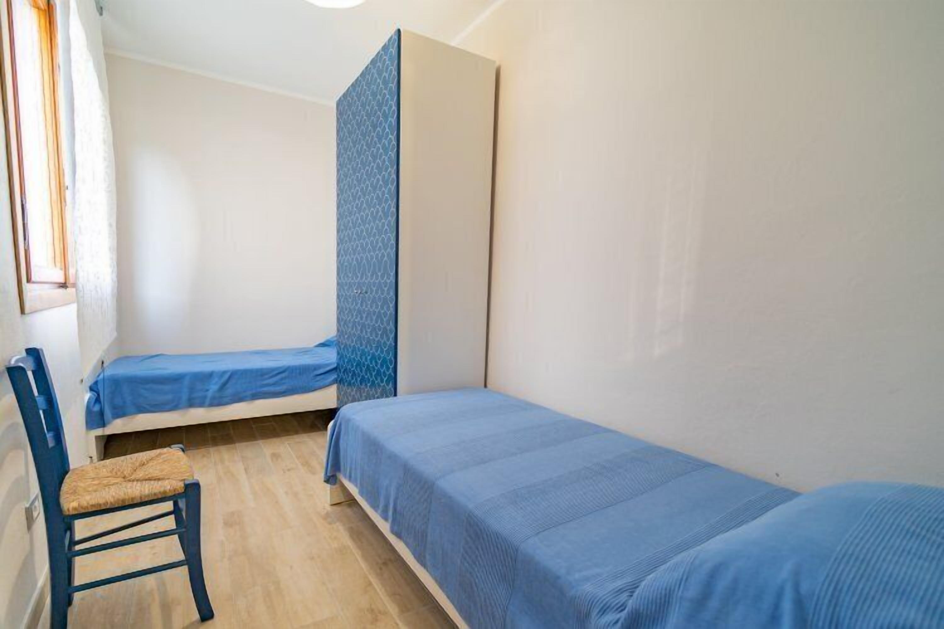 A small, single-room accommodation with two single beds, a chair, and a wardrobe.