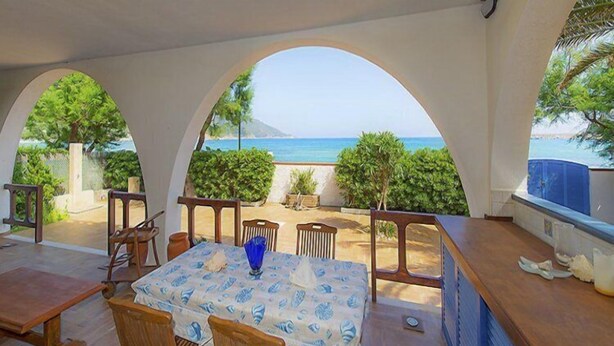 Un espace extérieur aménagé pour les repas, avec une table et des chaises, une vue sur la mer et un patio couvert agrémenté d’arcades.