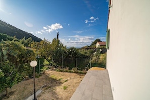 A courtyard with a paved walkway, a fenced area with vegetation, and a hillside in the background.