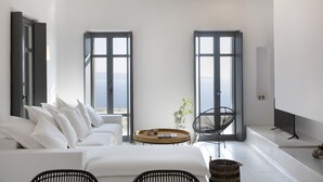 A modern room with a white sofa, a round wooden table, and a black chair. There are two large windows with black frames, and a flat-screen TV mounted on the wall. - House G in Kardiani Tinos (Tinos)