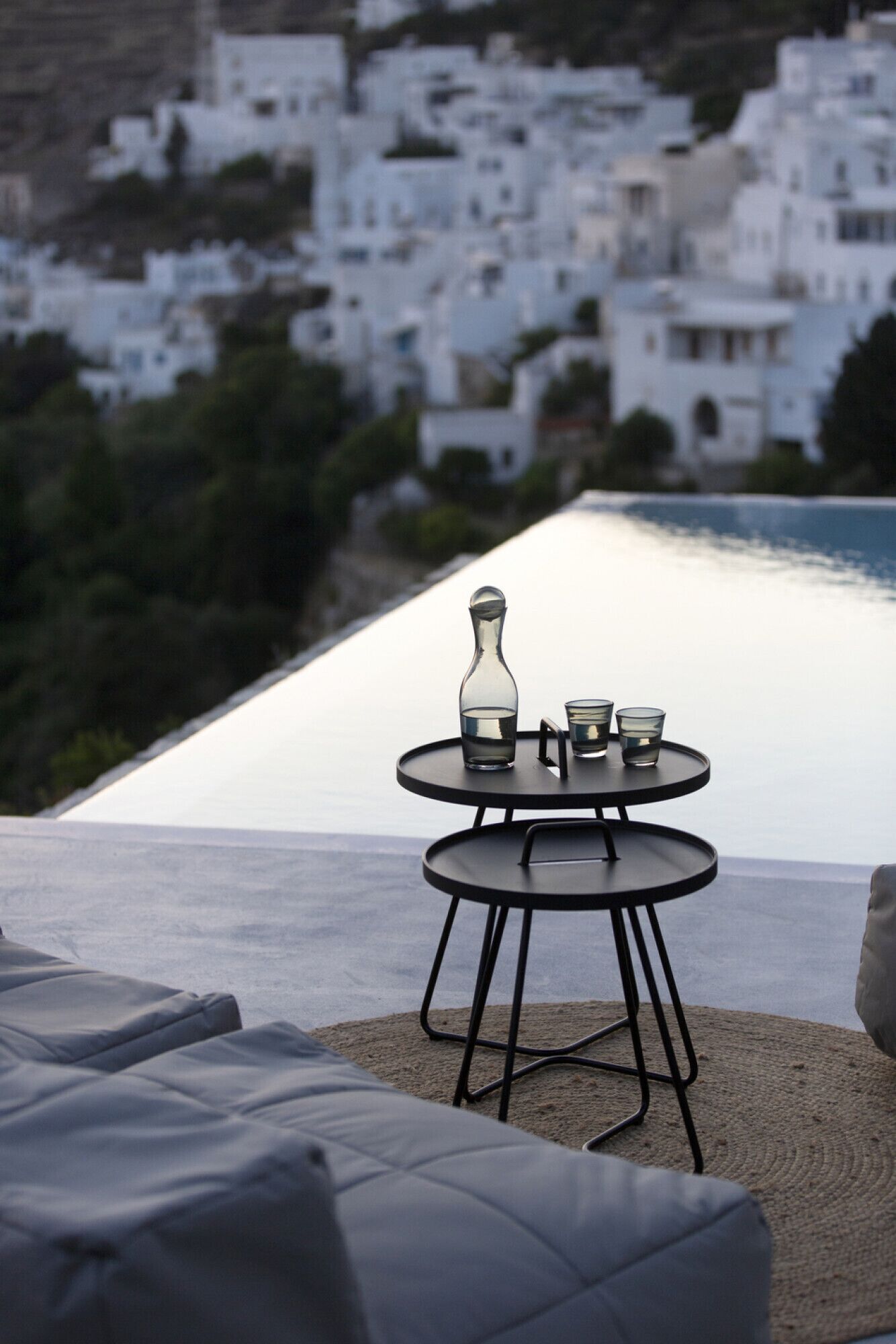 Une table d’extérieur à deux niveaux, sur laquelle sont posés une bouteille et des verres, le tout se détachant sur un arrière-plan composé d’immeubles blancs et d’une étendue d’eau.