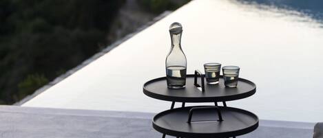 Une table d’extérieur à deux niveaux, sur laquelle sont posés une bouteille et des verres, le tout se détachant sur un arrière-plan composé d’immeubles blancs et d’une étendue d’eau.