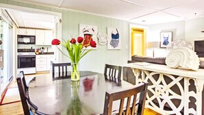 Un comedor con una mesa redonda, sillas, un florero con flores rojas y una cocina al fondo.